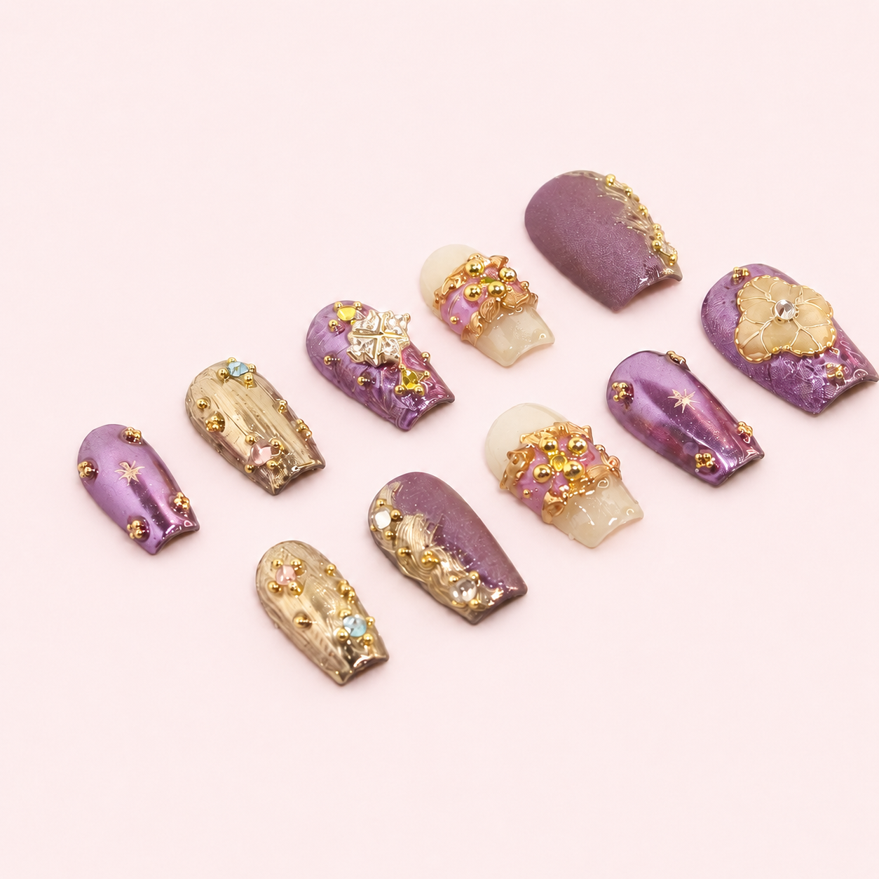 Set d’ongles press-on violets et dorés MB Beauty avec strass, fleurs 3D et finitions brillantes sur fond rose poudré.